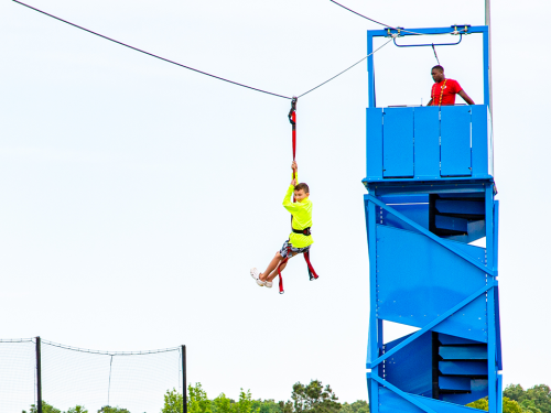 A child in a bright yellow shirt zip lines from a blue tower while an adult supervises from above.