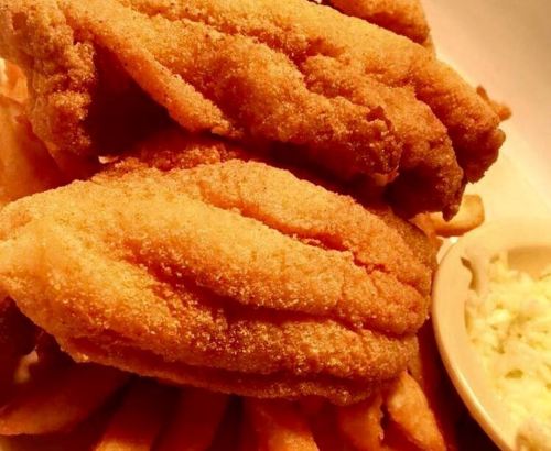A close-up of crispy fried fish fillets stacked on a bed of French fries, served with a side of coleslaw.