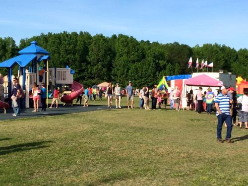 A lively outdoor scene with families enjoying a playground and colorful tents at a community event.