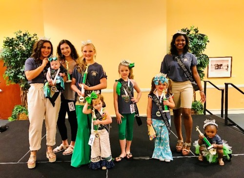 A group of young girls in pageant attire, holding trophies, posing on stage with smiles and sashes.