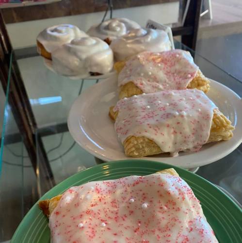 A display of pastries with white icing and pink sprinkles, including two cinnamon rolls in the background.