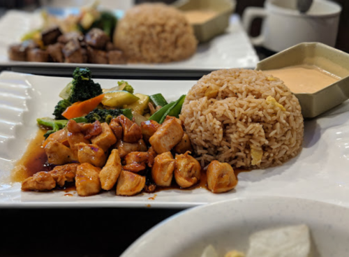 A plate of chicken stir-fry with vegetables, fried rice, and a small bowl of sauce.