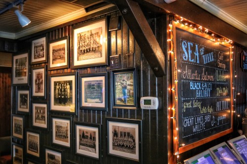 A cozy restaurant wall featuring framed historical photos and a chalkboard menu with decorative lights.