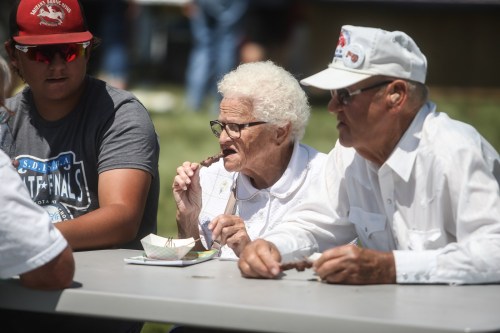 Three people sitting at a table outdoors, enjoying food and drinks on a sunny day. One woman has white hair and glasses.