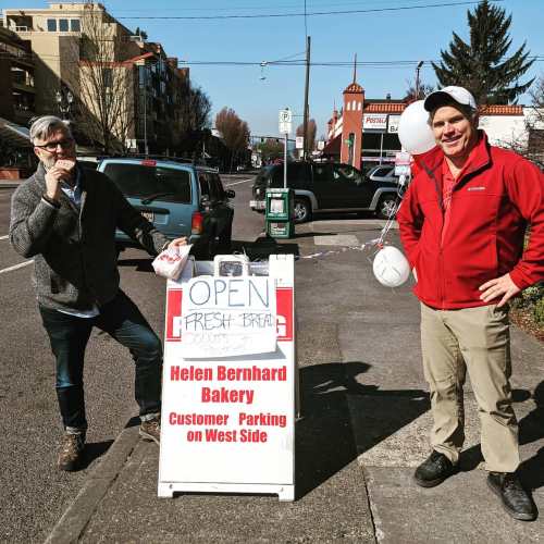 Two men stand beside a bakery sign that reads "Open" with balloons, indicating fresh bread and donuts available.