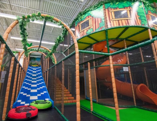 Colorful indoor play area featuring a slide, climbing structures, and a tunnel, surrounded by netting and greenery.