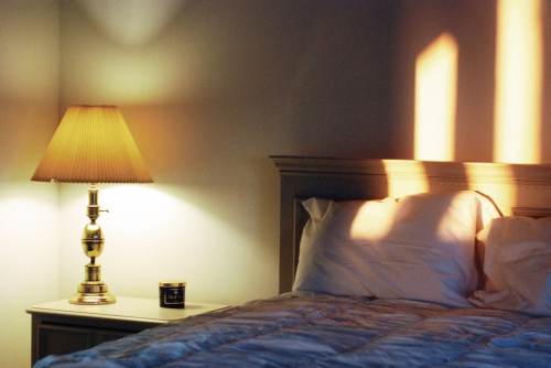 A softly lit bedroom with a lamp on a nightstand, casting warm shadows on the wall and a neatly made bed.