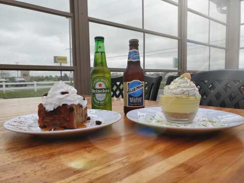 Two dessert plates on a wooden table: one with a cake topped with whipped cream, the other a layered cheesecake, with drinks.