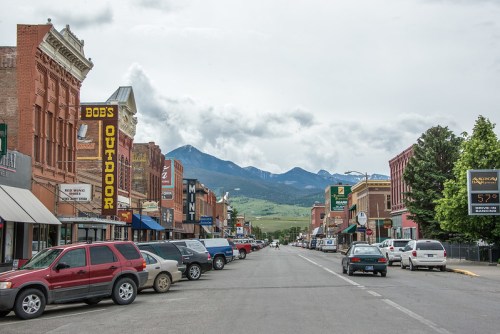 A street view of a small town with shops, parked cars, and mountains in the background under a cloudy sky.