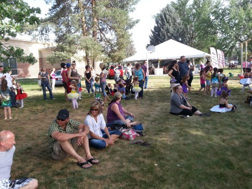 A crowd of people and children enjoying an outdoor event on a sunny day, with tents and trees in the background.