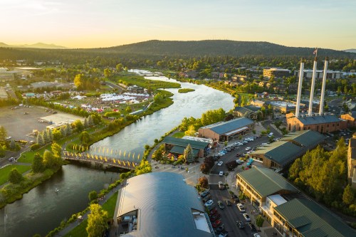 Aerial view of a river winding through a town, with green trees, buildings, and mountains in the background at sunset.
