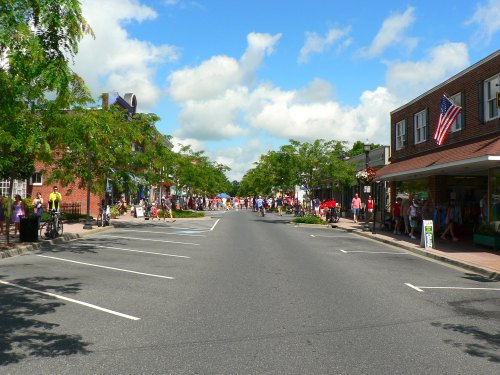 A lively street scene with shops, trees, and people walking under a blue sky with fluffy clouds.