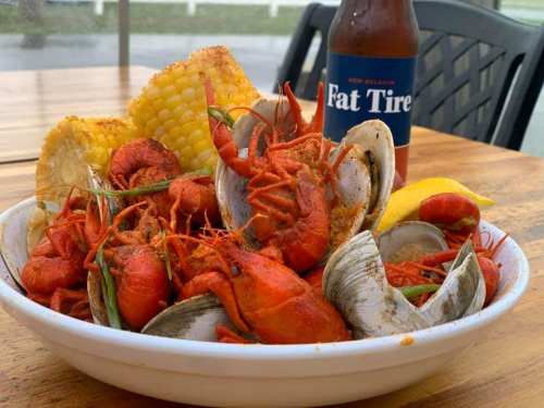 A bowl of spicy crawfish and clams, served with corn, lemon, and a bottle of Fat Tire beer on a wooden table.
