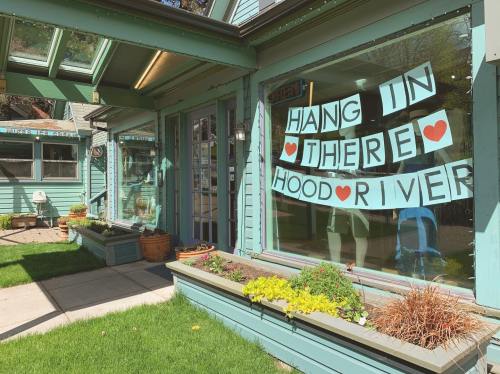 A storefront with a sign reading "HANG IN THERE HOOD RIVER" decorated with hearts, surrounded by greenery.