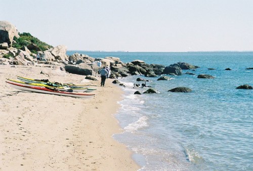 A sandy beach with kayaks on the shore, a person walking along the water's edge, and rocky formations in the background.
