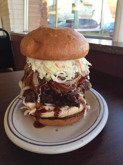 A towering burger with layers of meat, coleslaw, onions, and barbecue sauce, served on a plate in a diner setting.