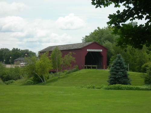 A red covered bridge sits on a grassy hill, surrounded by trees and under a cloudy sky.