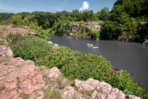 A serene river flows past rocky cliffs and lush greenery, with kayakers enjoying the calm water under a clear sky.