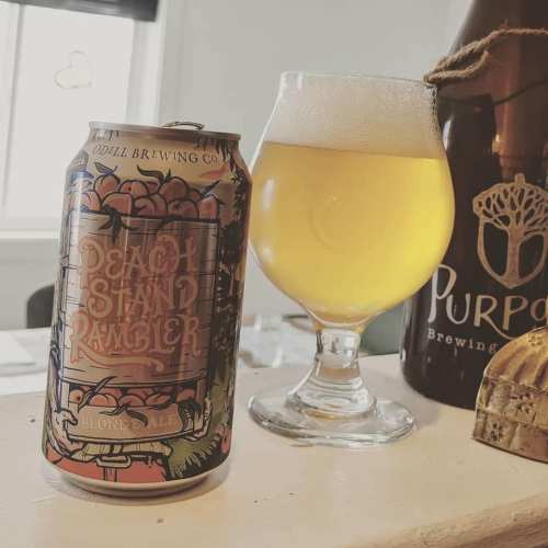 A can of Peach Stand Rambler blonde ale next to a glass of the beer, with a bottle labeled "Purpose Brewing" in the background.