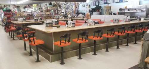 A spacious diner counter with orange and black stools, surrounded by shelves of various items and a kitchen area in the background.