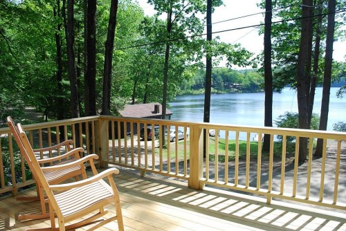 A serene lakeside view from a wooden deck with two rocking chairs, surrounded by lush green trees.