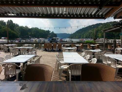 Outdoor dining area with white tables and chairs overlooking a marina filled with boats and lush green hills in the background.