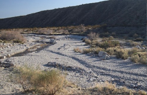A dry riverbed winds through a barren landscape, surrounded by sparse vegetation and rocky terrain under a clear sky.