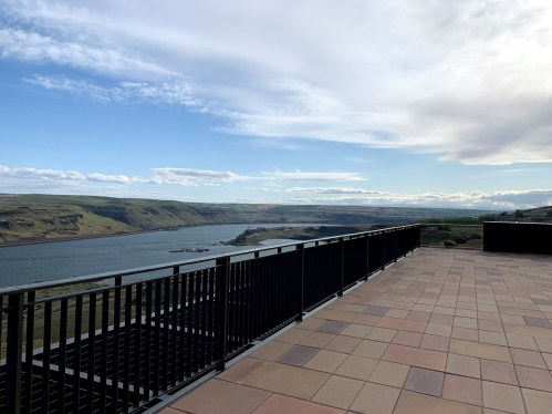 A scenic view of a river valley from a balcony, with rolling hills and a blue sky in the background.