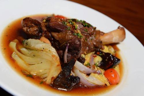 A plated dish featuring a tender meat shank with vegetables in a rich sauce, garnished with herbs.