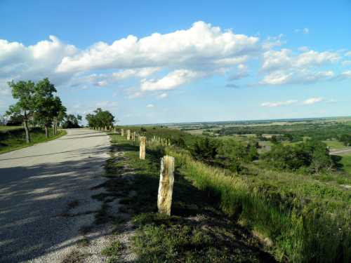 A scenic view of a gravel road lined with wooden posts, overlooking lush green fields and a blue sky with clouds.