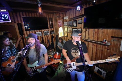 A live band performs in a cozy tavern, with musicians on guitars and vocals, and a drummer in the background.