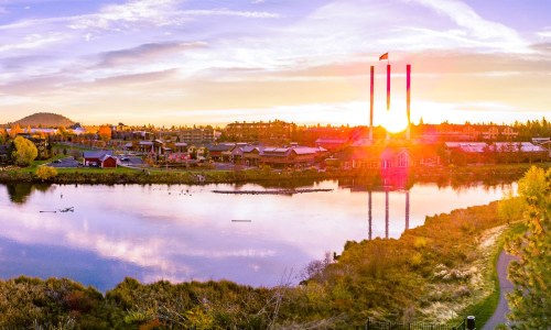 Sunset over a serene lake with reflections, featuring buildings and smokestacks in the background.