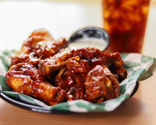 A plate of saucy chicken wings served with a side of dipping sauce, with a drink in the background.