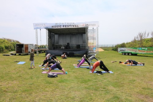 A group of people practicing yoga on mats outdoors, with a music festival stage in the background.