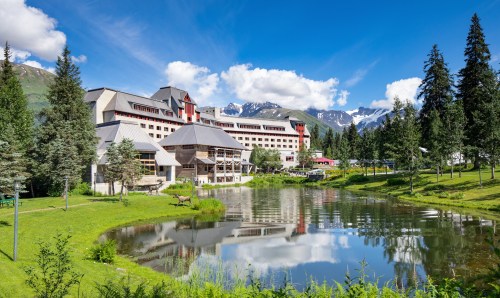 A scenic hotel by a pond, surrounded by lush greenery and mountains under a clear blue sky.