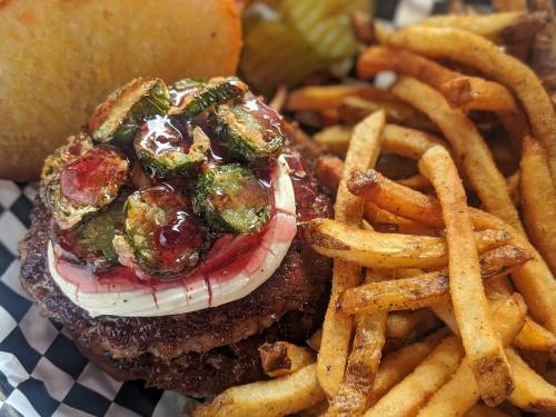 A juicy burger topped with crispy vegetables and sauce, served with golden fries and pickles on a checkered plate.