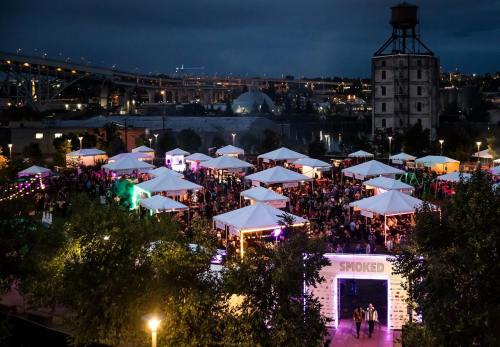 A vibrant outdoor event with tents, crowds, and colorful lights under a night sky, near a bridge and industrial buildings.