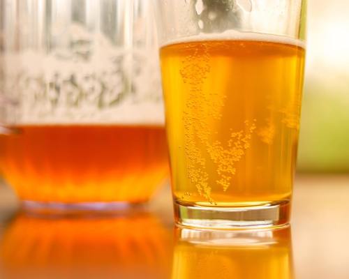 A close-up of a glass of golden beer with bubbles, next to a pitcher of beer in the background.