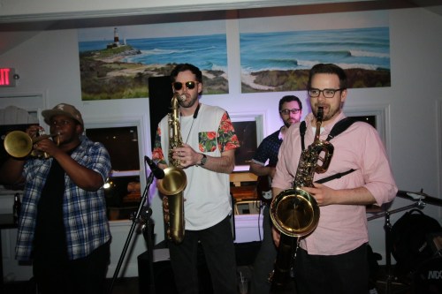 A band performs on stage with brass instruments in a lively setting, featuring a lighthouse mural in the background.