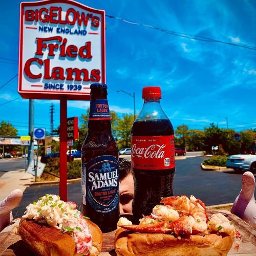 A person holds two lobster rolls, a Samuel Adams beer, and a Coca-Cola in front of Bigelow's Fried Clams sign.
