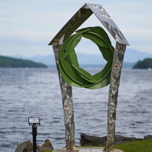 A metallic sculpture resembling a house frame with a green circular design, set by a lake with mountains in the background.