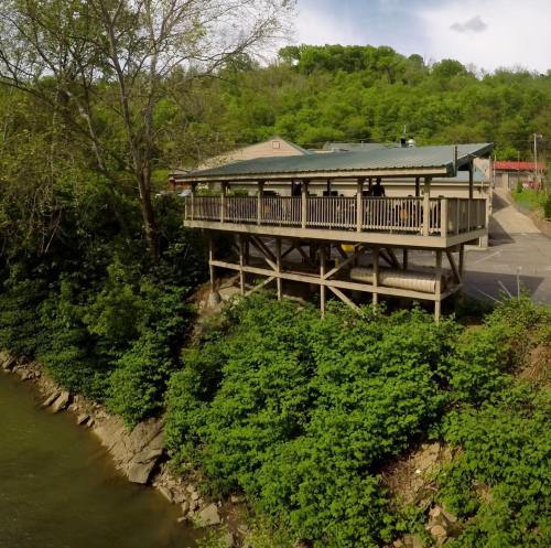 A wooden deck restaurant overlooks a river, surrounded by lush greenery and trees on a sunny day.