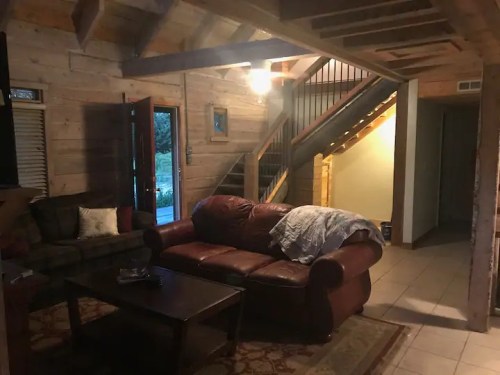 Cozy living room with a brown leather sofa, wooden walls, and a staircase in a rustic cabin setting.
