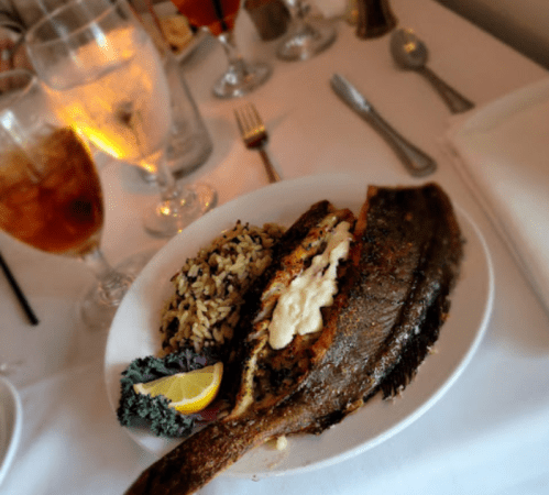 A plate of grilled fish with rice, a lemon wedge, and a side of greens, set on a dining table with drinks.