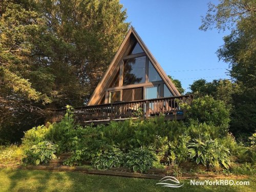 A charming A-frame cabin surrounded by lush greenery and trees, featuring large windows and a wooden deck.