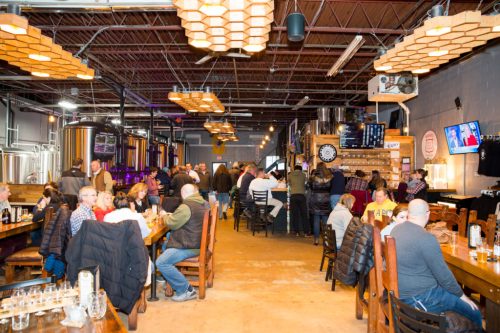 A lively brewery interior with patrons enjoying drinks, wooden tables, and large brewing equipment in the background.