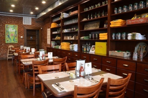 Cozy restaurant interior with wooden tables, shelves of drinks, and colorful towels against a brick wall.