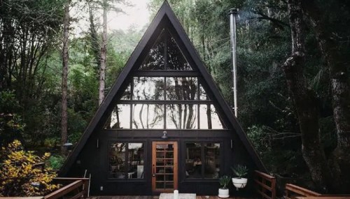A modern black A-frame cabin surrounded by lush trees, featuring large windows and a wooden deck.