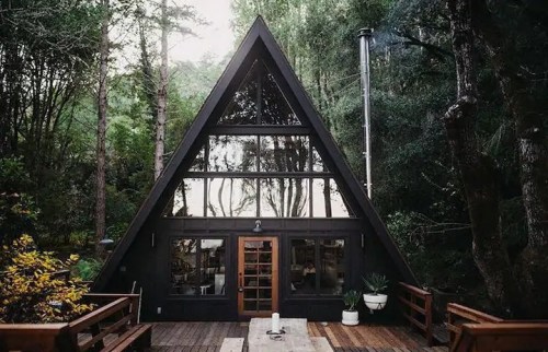 A modern A-frame cabin surrounded by trees, featuring large windows and a wooden deck.