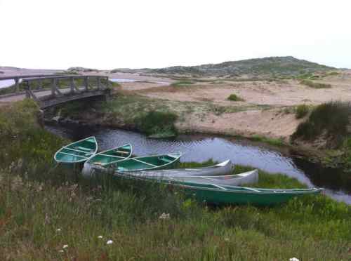 Canoes resting by a calm stream near a sandy landscape, with a bridge in the background and greenery surrounding.